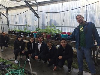 5 students sitting - 1 standing in greenhouse with plants on tables