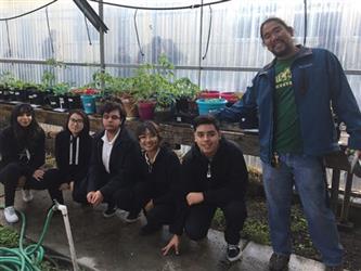 5 students sitting - 1 standing in greenhouse with plants on tables